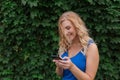 Beautiful young girl in a blue dress typing a message on the phone. Against the background of wild grapes, summer day. Copy space Royalty Free Stock Photo