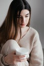 Beautiful young girl with brown hair in beige sweater looks away and holds a white cup in her hand. Portrait of girl drinking coff Royalty Free Stock Photo