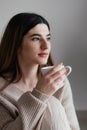 Beautiful young girl with brown hair in beige sweater looks away and holds a white cup in her hand. Portrait of girl drinking coff Royalty Free Stock Photo
