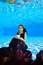 A beautiful young girl in a Burgundy dress sits and poses underwater at the bottom of the pool, holds a white Cup in her hand Royalty Free Stock Photo