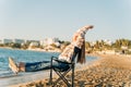 Beautiful young girl in cozy sweater stretching while sitting on foldable chair on winter beach sand. Woman enjoying Royalty Free Stock Photo