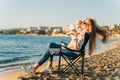 Beautiful young girl in cozy sweater and sunglasses stretching while sitting on foldable chair on winter beach sand Royalty Free Stock Photo