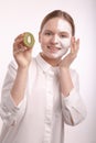 Beautiful young girl with a cream mask on her face holding a kiwi.