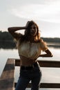 Beautiful young girl with curly hairstyle in fashionable summer denim clothes resting and poses near the lake at sunset Royalty Free Stock Photo