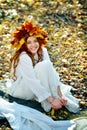 A beautiful young girl dressed in white, wearing a wreath of yellow leaves, sitting on fall leaves on a Sunny day Royalty Free Stock Photo