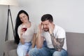 Beautiful young girl drinking wine sitting on the couch, and her boyfriend is sitting frustrated. He has a headache Royalty Free Stock Photo