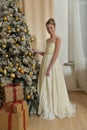 Beautiful young girl with evening hairstyle in a white dress by the Christmas tree