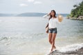 A beautiful young girl in a hat and with a backpack playfully walks by the water. A warm summer day is a great time for Royalty Free Stock Photo
