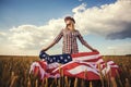 Beautiful young girl holding an American flag on the wind in a field of wheat. Summer landscape against the blue sky Royalty Free Stock Photo