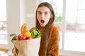 Beautiful young girl holding paper bag of fresh groceries scared in shock with a surprise face, afraid and excited with fear