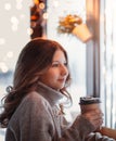 Beautiful young girl holds a paper cup of coffee and looks out the window of the cafe Royalty Free Stock Photo