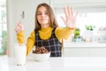 Beautiful young girl kid eating chocolate cereals and glass of milk for breakfast with open hand doing stop sign with serious and Royalty Free Stock Photo