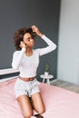 Beautiful young girl listening music in earphones on pink bed, touching her brown curly hair. Room with gray wall on Royalty Free Stock Photo