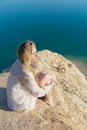 Beautiful young girl with long blond hair in a white dress sitting on the beach, the lake on a bright sunny day Royalty Free Stock Photo