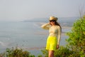 Beautiful young girl with long hair in straw hat, dark glasses and short yellow skirt stands on top against coastline and beach