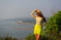 Beautiful young girl with long hair in straw hat, dark glasses and short yellow skirt stands on top against coastline and beach in