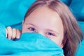 Beautiful young girl lying down in the bed and sleeping. Teen girl with open eyes covers her face with blue blanket in the morning
