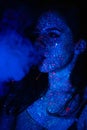 A beautiful young girl with makeup from ultraviolet paints smokes. Releases smoke rings from the mouth. Holiday of