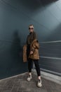 Beautiful young girl model in a fashionable leather jacket with black jeans and sneakers in sunglasses near a metal modern wall in Royalty Free Stock Photo