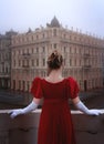 Beautiful young girl in a nineteenth century red dress stands on a balcony in front of a beautiful building