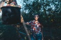 Beautiful young girl, in a red plaid shirt, and jeans, leaned on a ladder with a bucket, autumn harvest, apples, Royalty Free Stock Photo