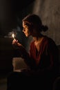 Beautiful young girl in red shirt lights up her pretty face with match sitting in dark room and holding matchbox in hand