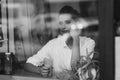 A beautiful young girl sits in a cafe behind a glass with a cup of coffee and looks thoughtfully out the window. Black and white Royalty Free Stock Photo