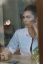 A beautiful young girl sits in a cafe behind a glass with a cup of coffee and looks thoughtfully out the window. Photo through Royalty Free Stock Photo