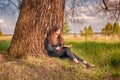 A beautiful young girl is sitting in nature under a large old tree and reading a book in the open air on a Sunny day. Royalty Free Stock Photo