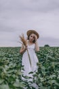 Beautiful young girl in white dress and straw hat in green field hugging bouquet of dried herbs