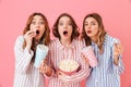 Beautiful young girls 20s wearing colorful striped pyjamas having fun and eating pop corn during watching thrilling and exciting