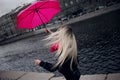 Beautiful young and happy blond woman in a bright pink scarf, rubber boots and umbrella walking in a rainy city. Royalty Free Stock Photo