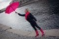 Beautiful young and happy blond woman in a bright pink scarf, rubber boots and umbrella walking in a rainy city. Royalty Free Stock Photo