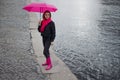 Beautiful young and happy blond woman in a bright pink scarf, rubber boots and umbrella walking in a rainy city. Royalty Free Stock Photo