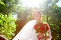 Beautiful young happy bride standing near green trees in a park and holding a beautiful bouquet. Royalty Free Stock Photo