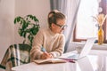 Beautiful young happy girl student with glasses and a warm siver studying at home online with a laptop writing in a notebook