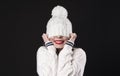 Beautiful young happy smiling girl hiding her eyes under trendy white big loop knitted beanie hat. Model wearing winter sweater. Royalty Free Stock Photo