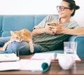 Beautiful young happy woman in glasses uses a smartphone on the couch, a cute red cat lap Royalty Free Stock Photo