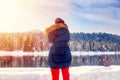 Beautiful young hiker girl in down jacket and red pants photographing winter landscape with river, mountains and forest at sunset Royalty Free Stock Photo