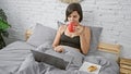 Beautiful young hispanic woman chilling in her bedroom, having a relaxed morning with breakfast in bed, using laptop and enjoying Royalty Free Stock Photo