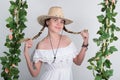 Beautiful young leggy blonde in a little white dress and white cowboy hat on a swing, wooden swing suspended from a rope Royalty Free Stock Photo