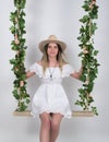 Beautiful young leggy blonde in a little white dress and white cowboy hat on a swing, wooden swing suspended from a rope Royalty Free Stock Photo