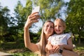 Beautiful young mother and daughter with blonde hair using mobile phone outdoor. Stylish girls making selfie in the park. People