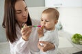 Beautiful young mother feeding little baby boy with spoon while he already eating cookie. Child looks at spoon with Royalty Free Stock Photo