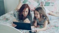 Beautiful young mother and her cute daughter in pajamas laughing and looking in digital tablet while lying on bed at Royalty Free Stock Photo