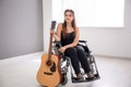 Beautiful young musician with guitar sitting in wheelchair indoors Royalty Free Stock Photo