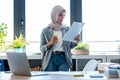 Beautiful young muslim business woman wearing hijab using her smart phone while holding papers looking through the window in the Royalty Free Stock Photo