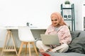 A beautiful young Muslim woman wearing casual clothing is using a laptop and smiling while working at home, sitting on a Royalty Free Stock Photo