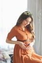 A beautiful young pregnant girl with a wreath of flowers on her head in a brown dress with lace is sitting near the window and Royalty Free Stock Photo