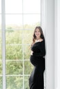 beautiful young pregnant woman in dark black dress stands near the window Royalty Free Stock Photo